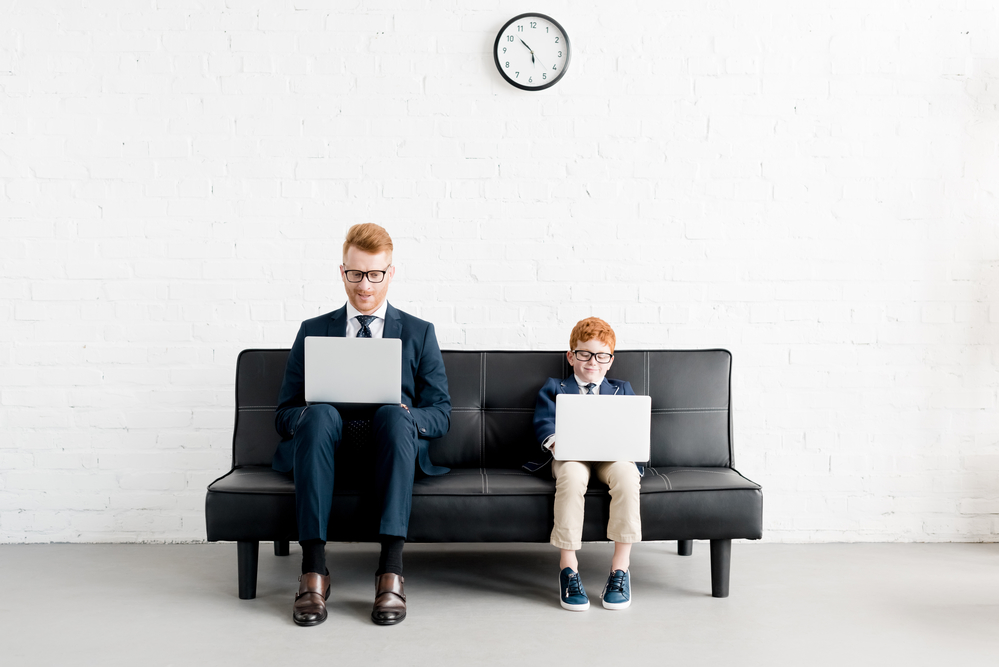 Photo of a man and a child sitting on a black sofa, both using laptop laptops